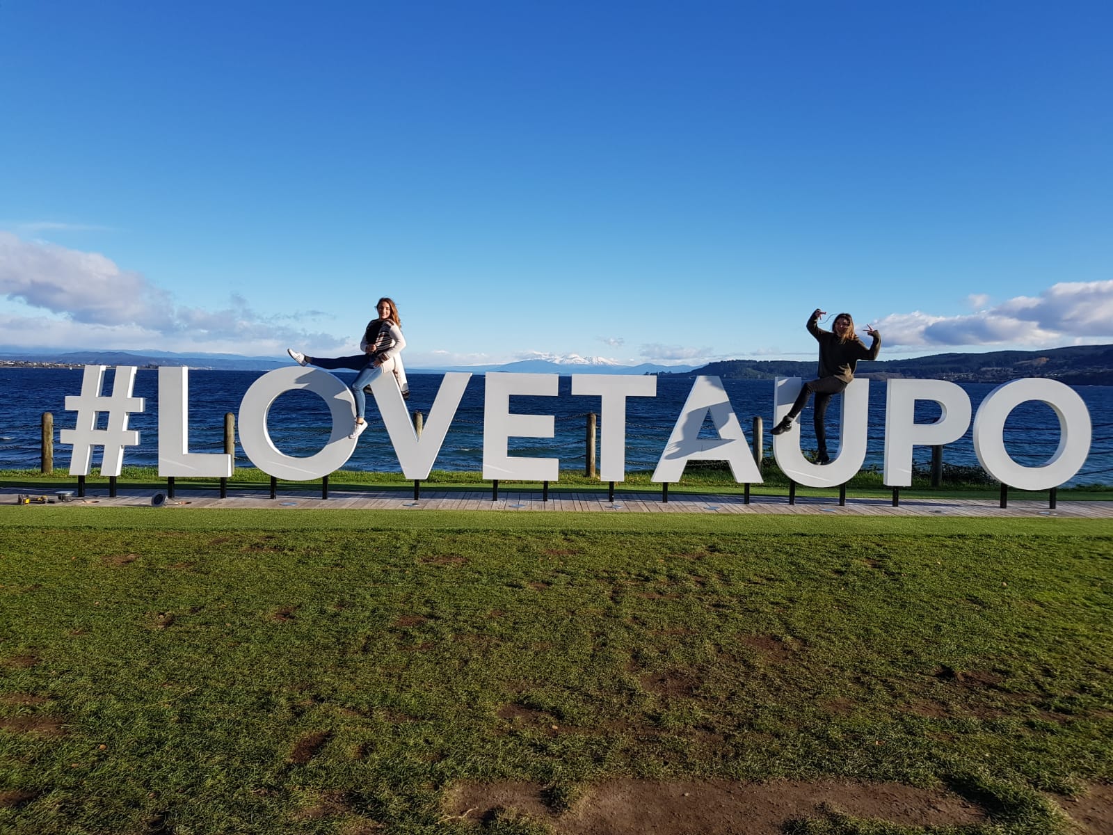 People posing by the #LOVETAUPO sign on the grassy shores of Lake Taupō, New Zealand.